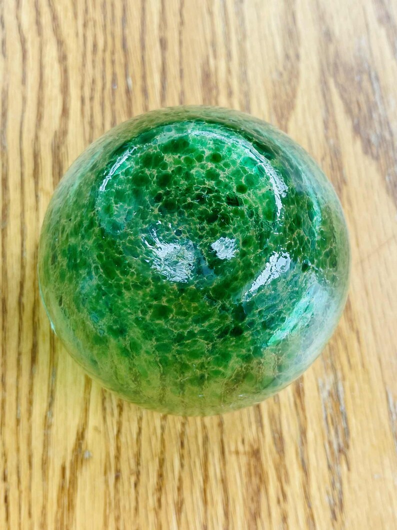 Puede incluir: Un objeto decorativo redondo de vidrio verde con una superficie texturizada. El objeto es de un tono verde vibrante y parece estar hecho de vidrio. El objeto est&aacute; sobre una superficie de madera.