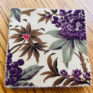May include: Square fabric with a floral pattern. The design features large purple flowers, brown leaves, and red accents on a cream background. The edges are finished with a scalloped trim.