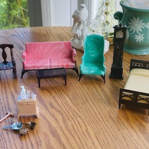 May include: A collection of miniature furniture pieces, including a pink sofa, a green armchair, a white armchair, a wooden bed, a wooden dresser, a wooden table, and several chairs. The furniture is made of plastic and wood and is painted in various colors.