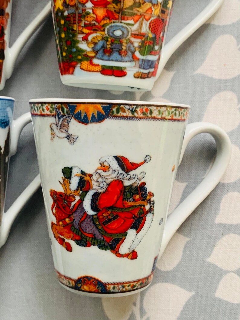 May include: White ceramic mug with a Christmas scene featuring Santa Claus riding a reindeer. The mug has a blue and gold trim around the top.