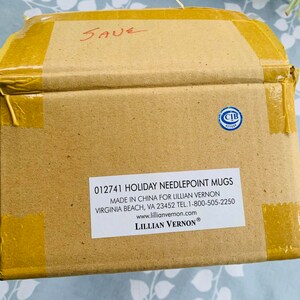 May include: A brown cardboard box with a label that reads "012741 HOLIDAY NEEDLEPOINT MUGS MADE IN CHINA FOR LILLIAN VERNON VIRGINIA BEACH, VA 23452 TEL. 1-800-505-2250 www.lillianvernon.com LILLIAN VERNON&reg;". The box is taped shut with yellow tape.