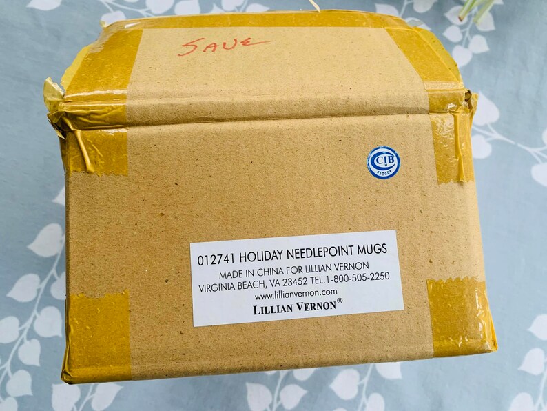 May include: A brown cardboard box with a label that reads "012741 HOLIDAY NEEDLEPOINT MUGS MADE IN CHINA FOR LILLIAN VERNON VIRGINIA BEACH, VA 23452 TEL. 1-800-505-2250 www.lillianvernon.com LILLIAN VERNON&reg;". The box is taped shut with yellow tape.
