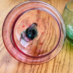 Puede incluir: Primer plano de un vaso transparente te&ntilde;ido de rosa con un centro oscuro. El vaso tiene forma circular con un borde grueso. El fondo muestra una superficie de madera y un vaso verde.