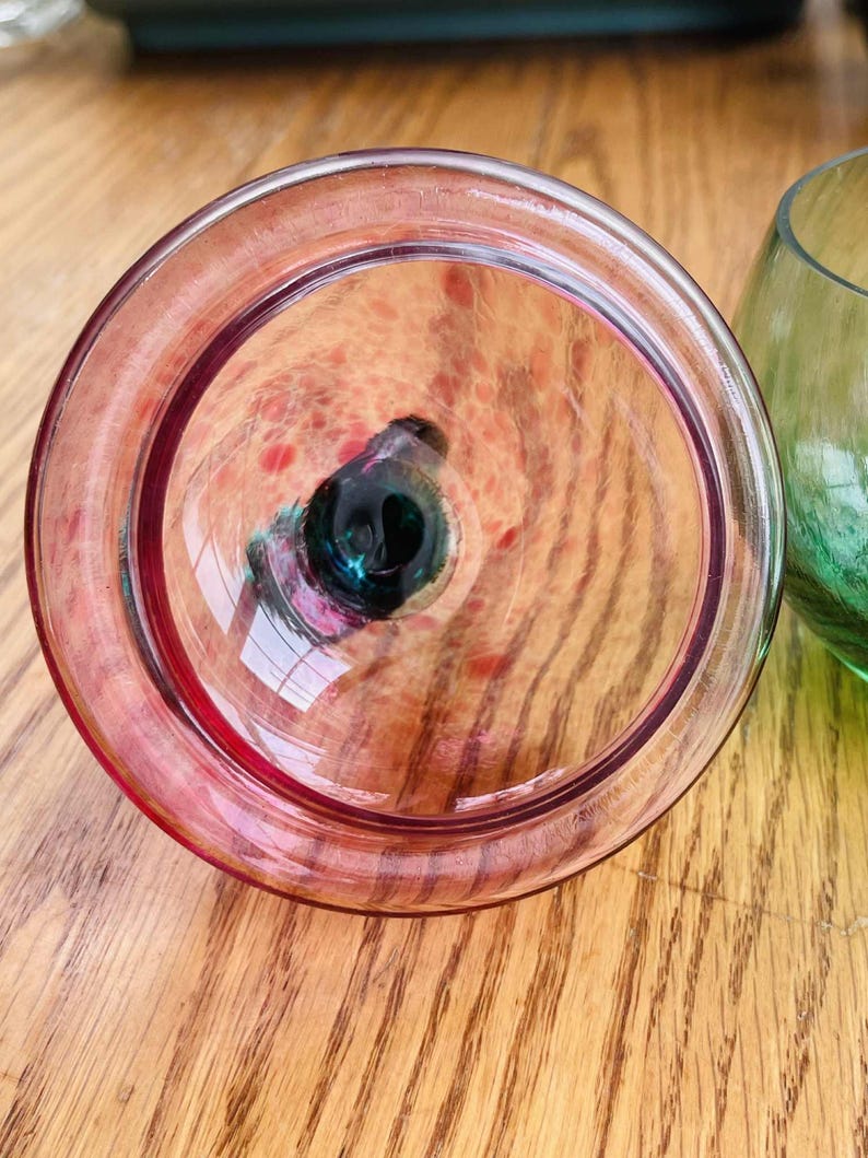 Puede incluir: Primer plano de un vaso transparente te&ntilde;ido de rosa con un centro oscuro. El vaso tiene forma circular con un borde grueso. El fondo muestra una superficie de madera y un vaso verde.