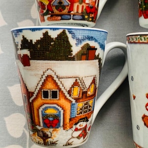 May include: A white ceramic mug with a winter scene featuring a house with a red bow on the door, a snowman, and trees. The mug has a blue rim and a white handle.