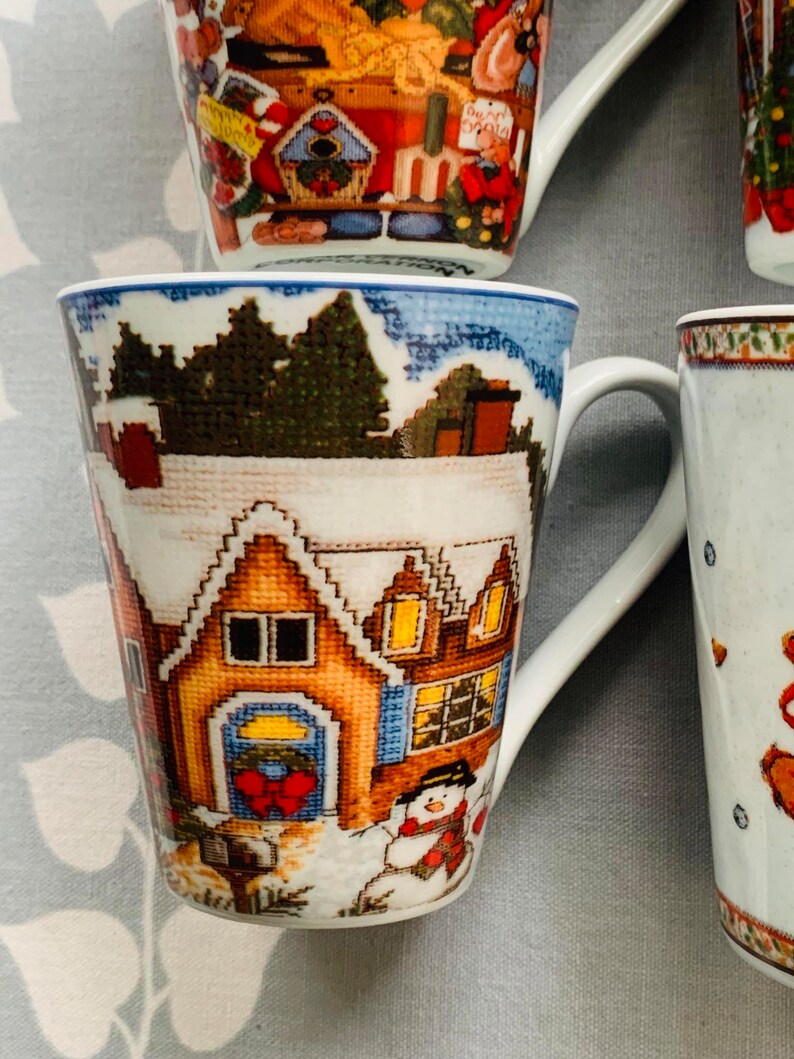 May include: A white ceramic mug with a winter scene featuring a house with a red bow on the door, a snowman, and trees. The mug has a blue rim and a white handle.