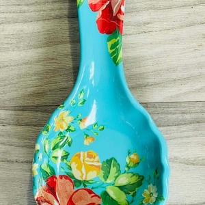 May include: A turquoise ceramic spoon rest with a floral design. The spoon rest features a wide, shallow bowl and a long handle. The floral pattern includes red, yellow, and orange flowers with green leaves.