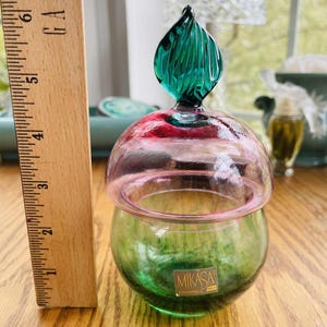 Puede incluir: Un tarro de vidrio decorativo con base verde y tapa rosa y roja. El tarro tiene un tap&oacute;n verde en forma de hoja. El tarro est&aacute; etiquetado "MIKASA CZECH REPUBLIC". Una regla de madera est&aacute; al lado del tarro.