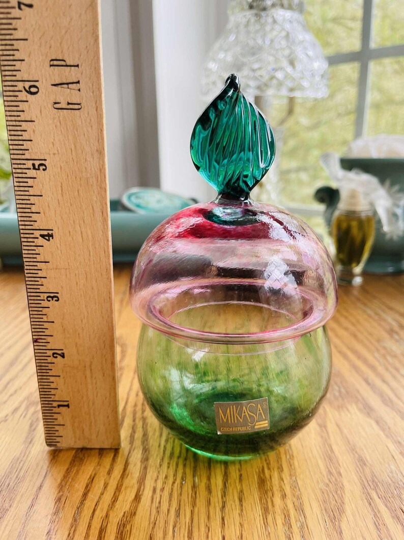 Puede incluir: Un tarro de vidrio decorativo con base verde y tapa rosa y roja. El tarro tiene un tap&oacute;n verde en forma de hoja. El tarro est&aacute; etiquetado "MIKASA CZECH REPUBLIC". Una regla de madera est&aacute; al lado del tarro.