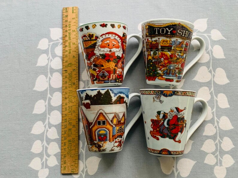 May include: Four ceramic mugs with Christmas-themed designs. The mugs feature Santa Claus, a toy shop, a winter village, and Santa Claus riding a reindeer. The mugs are white with colorful designs.