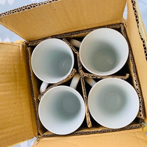 May include: A set of four white ceramic mugs in a brown cardboard box. The mugs are arranged in a square pattern with the handles facing outward.