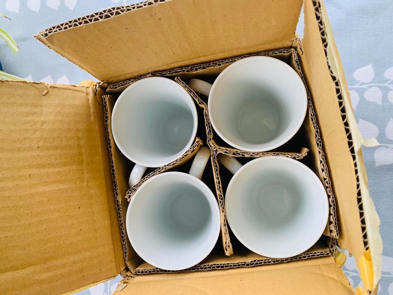 May include: A set of four white ceramic mugs in a brown cardboard box. The mugs are arranged in a square pattern with the handles facing outward.