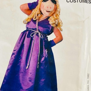 May include: A vintage Miss Piggy costume from Jim Henson's Muppet Costumes. The costume features a purple satin dress with a rope belt, matching gloves, and a Miss Piggy mask with blonde curly hair. The image is from 1982.