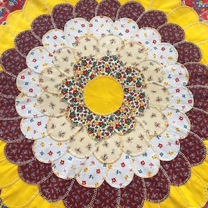 May include: A circular fabric mat featuring a layered petal design in a sunflower pattern. The center is a solid yellow circle surrounded by concentric rings of fabric petals in floral prints, solid yellow, and dark maroon with stitched edges.