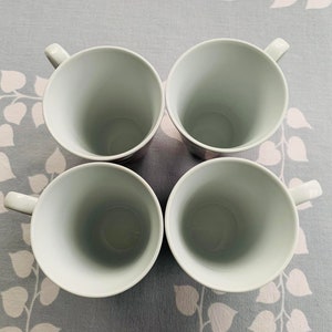 May include: Four white ceramic mugs with rounded handles. The mugs are arranged in a square formation on a light gray background with a floral pattern.
