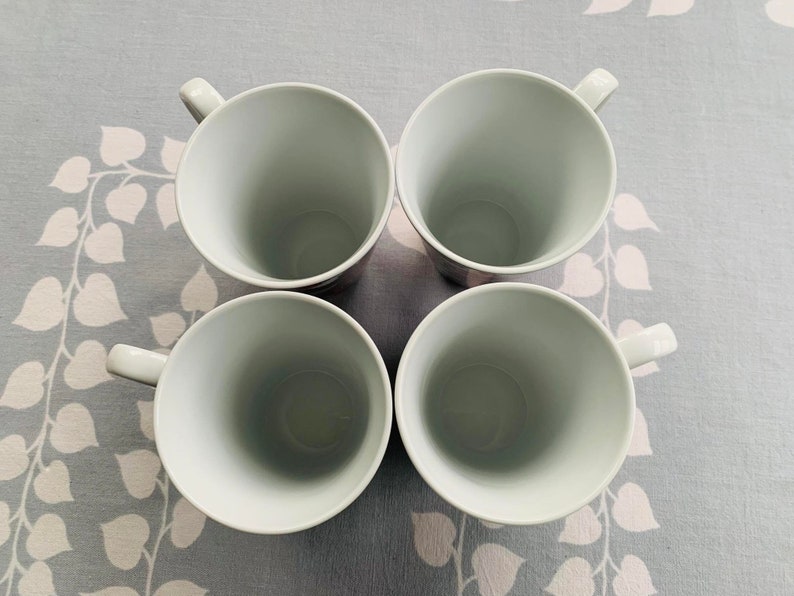 May include: Four white ceramic mugs with rounded handles. The mugs are arranged in a square formation on a light gray background with a floral pattern.