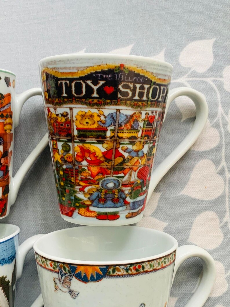 May include: A set of four white ceramic mugs with colorful holiday designs. The mugs feature a toy shop, a winter scene, a bird, and a snowman. The mugs have a handle on the right side.