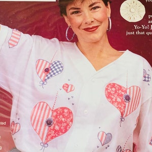 May include: A white shirt with a red, white, and blue heart design. The shirt has a button-up collar and short sleeves. The model is wearing a blue baseball cap with a red, white, and blue flower design.