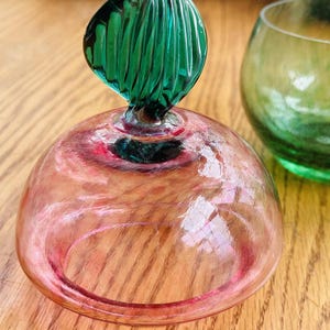 Puede incluir: Un tap&oacute;n de botella decorativo de vidrio con una c&uacute;pula rosa y un asa en forma de hoja verde. El tap&oacute;n est&aacute; junto a un vaso verde. El vaso est&aacute; sobre una superficie de madera.