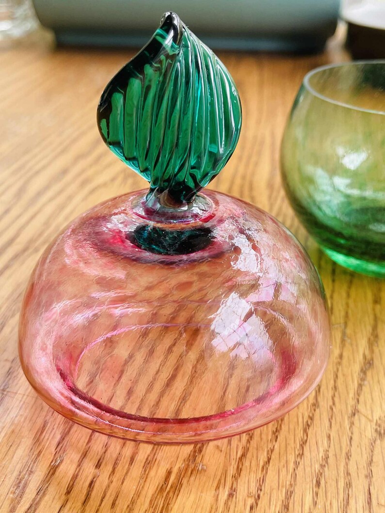 Puede incluir: Un tap&oacute;n de botella decorativo de vidrio con una c&uacute;pula rosa y un asa en forma de hoja verde. El tap&oacute;n est&aacute; junto a un vaso verde. El vaso est&aacute; sobre una superficie de madera.