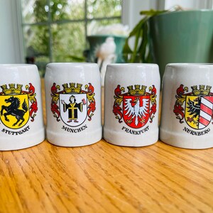 May include: Four small white ceramic mugs with colorful crests and city names. The mugs are labeled "Stuttgart", "Munchen", "Frankfurt", and "Nurnberg".