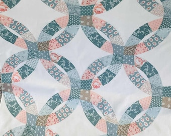 May include: A patchwork quilt with a double wedding ring design. The quilt features a repeating pattern of interlocking rings in shades of pink, teal, grey and brown, set against a white backdrop. The quilt is likely made of cotton fabric.