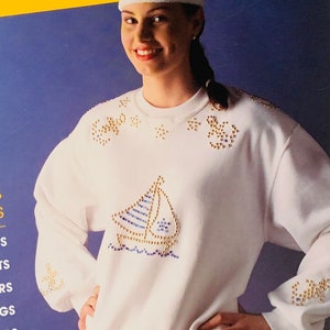 May include: White sweatshirt with a sailboat design made of gold and blue rhinestones. The sweatshirt has a white headband and a ponytail.