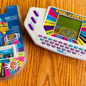 May include: A white Tiger Electronics Wheel of Fortune handheld game console with purple buttons and a QWERTY keyboard, alongside a sealed blister pack containing a Wheel of Fortune game cartridge featuring a colorful prize wheel graphic.