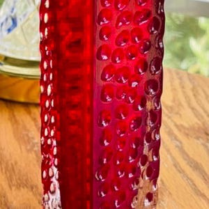 May include: A tall, square-shaped red glass vase featuring a textured surface with numerous small, circular indentations. The base of the vase transitions into a clear, thick glass bottom, providing a contrast to the vibrant red upper section.