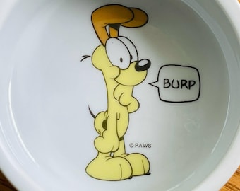 Puede incluir: Cuenco de cerámica blanco con un personaje de perro de dibujos animados amarillo y marrón. El perro tiene ojos grandes y un bocadillo que dice "BURP". El cuenco tiene un diseño sencillo, adecuado para comida o agua para mascotas.