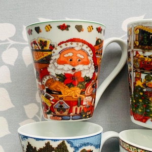 May include: Four ceramic mugs with Christmas-themed designs. One mug features a Santa Claus illustration, another has a winter village scene, and two others have a Christmas tree and a snowman.