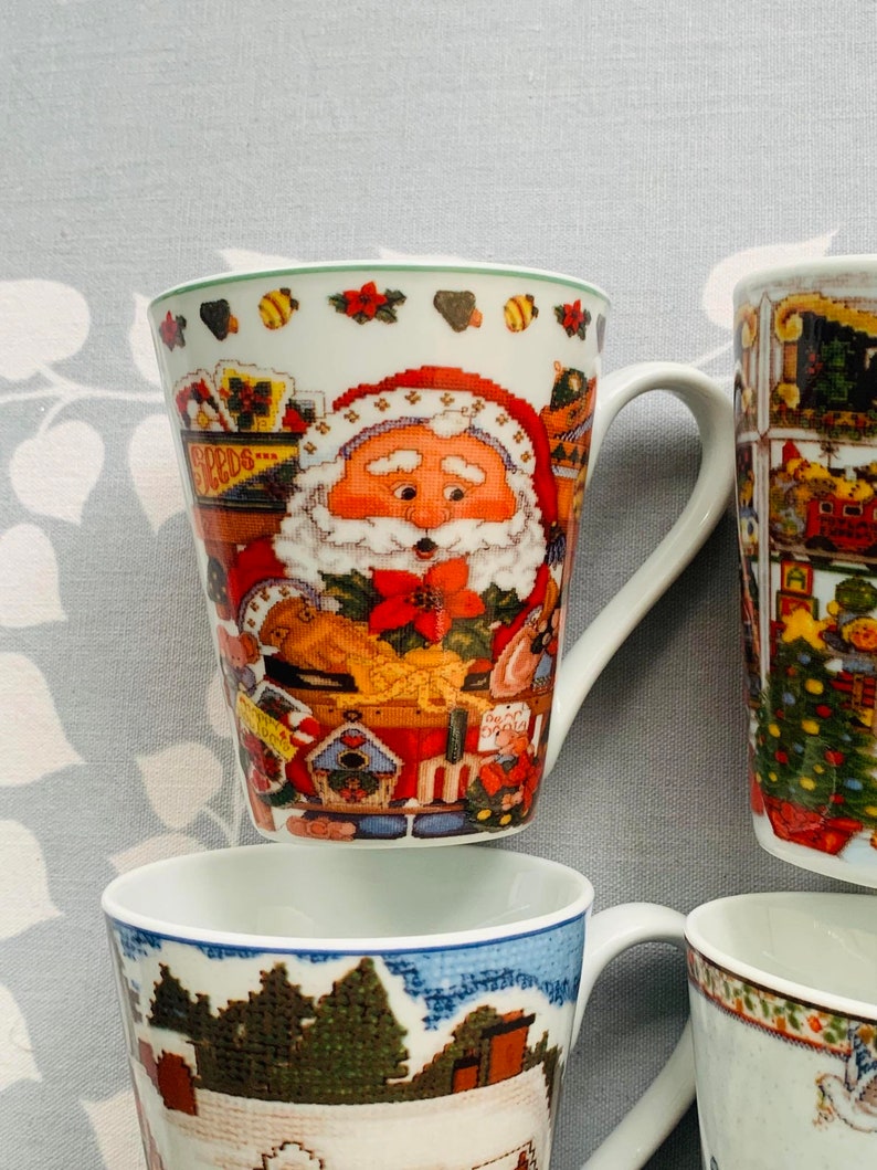 May include: Four ceramic mugs with Christmas-themed designs. One mug features a Santa Claus illustration, another has a winter village scene, and two others have a Christmas tree and a snowman.