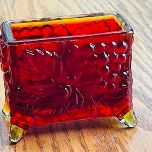 May include: A rectangular ruby red glass planter featuring a raised relief pattern of grape clusters and leaves on all sides. The vessel is supported by four small, clear glass feet at the corners, creating a distinct contrast with the deep red body.