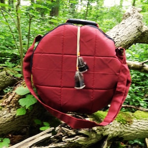 Può includere: Una borsa a tracolla trapuntata bordeaux con manico e tracolla neri. La borsa ha un ciondolo decorativo con piume. La borsa è rotonda e presenta un motivo a griglia cucita. Lo sfondo è una foresta.