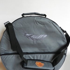 Könnte beinhalten: Eine kreisförmige graue Tragetasche mit einem gestickten Federdesign auf der Vorderseite. Die Tasche verfügt über einen schwarzen Tragegriff, einen verstellbaren schwarzen Schultergurt mit Polsterung und ein kleines orangefarbenes Logo.