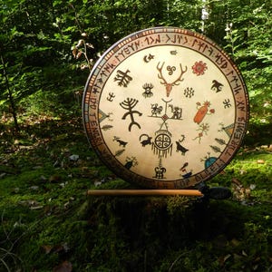 Op de afbeelding: Een ronde frametrommel met een lichtgekleurd vel voorzien van diverse zwarte en gekleurde symbolen, waaronder een hertenkop, geometrische vormen en figuren. Een rand met tekst omcirkelt de trommel. Een houten drumstok ligt horizontaal ervoor.