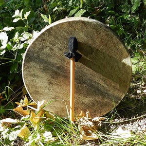 Op de afbeelding: Een ronde frametrommel met een gestructureerd, lichtbruin dierenhuidvel, rechtop geplaatst. Een houten klopper met een zwarte, afgeronde, met stof beklede kop en een kleine zwarte stoffen ruche aan de basis van de kop rust tegen de trommel.