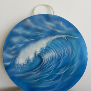 Könnte beinhalten: Ein kreisförmiger Wandbehang mit einem Airbrush-Gemälde einer brechenden Meereswelle. Das Kunstwerk zeigt Schattierungen von tiefem und hellem Blau mit weißem Schaum an der Wellenkrone. Eine kleine weiße Aufhängeschlaufe ist oben angebracht.