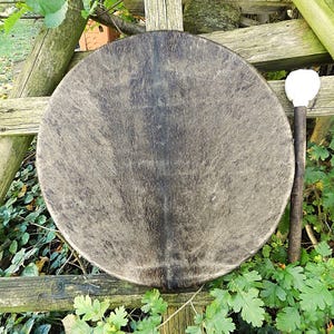 Op de afbeelding: Een ronde frametrommel met een donkere, getextureerde dierenhuid met natuurlijke bruine en zwarte vachtpatronen. De trommel is voorzien van een houten handvat en een bijpassende klopper met een witte, zachte, ronde kop op een houten stok.