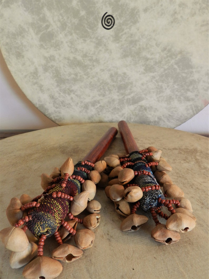 Puede incluir: Dos instrumentos de percusi&oacute;n, probablemente sonajeros o maracas, con mangos de madera y racimos de vainas. Los mangos son de color marr&oacute;n oscuro y las vainas de color marr&oacute;n claro. Descansan sobre un tambor con un s&iacute;mbolo de espiral.