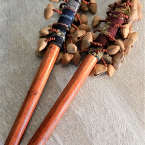 Puede incluir: Dos instrumentos de percusi&oacute;n de madera, cada uno con un mango largo y pulido y una empu&ntilde;adura de tela tejida adornada con cuentas. La parte superior de cada instrumento est&aacute; cubierta de vainas de semillas. Los instrumentos descansan sobre una superficie clara y texturizada.