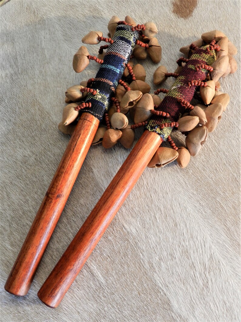 Puede incluir: Dos instrumentos de percusi&oacute;n de madera, cada uno con un mango largo y pulido y una empu&ntilde;adura de tela tejida adornada con cuentas. La parte superior de cada instrumento est&aacute; cubierta de vainas de semillas. Los instrumentos descansan sobre una superficie clara y texturizada.