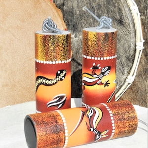 Pode incluir: Três instrumentos de percussão de madeira com desenhos pintados de lagartos e cobras. Os instrumentos são laranja, marrom e branco com detalhes em preto. Cada instrumento tem um laço de metal na parte superior.