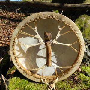 Op de afbeelding: Een ronde, handgemaakte trommel met een natuurlijke huidkop en een houten frame. De trommel heeft een gesneden berenhandvat en is versierd met natuurlijke vezels. De trommel rust op een mosachtige ondergrond in een bosrijke omgeving.
