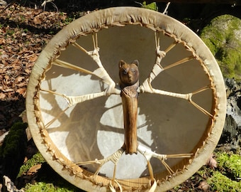 May include: A round, handcrafted drum with a natural hide head and a wooden frame. The drum features a carved bear handle and is decorated with natural fibers. The drum is resting on a mossy surface in a forest setting.
