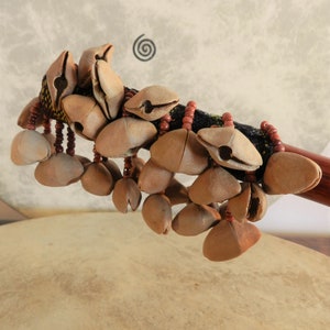 Puede incluir: Primer plano de un instrumento de percusi&oacute;n, probablemente una sonaja o un agitador. El instrumento presenta numerosas vainas de semillas de color marr&oacute;n claro unidas a un mango tejido oscuro con cuentas rojas. El fondo muestra un parche de tambor y un dise&ntilde;o en espiral.