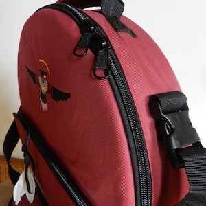Pode incluir: Uma mochila bordô com fecho de correr preto, alça superior e uma alça preta ajustável com fivela de plástico. A parte frontal apresenta um gráfico de um pássaro com um halo amarelo e um design de semicírculo laranja na base.