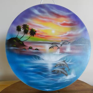 Pode incluir: Uma tábua de corte redonda de vidro com uma cena de pôr do sol tropical feita com aerógrafo, apresentando palmeiras numa pequena ilha, águas oceânicas azuis e quatro golfinhos a saltar acima da superfície. Inclui uma pega de plástico.