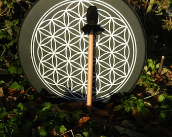 May include: A round black drum featuring a white geometric flower of life pattern centred on its surface, accompanied by a wooden mallet with a black felt head positioned vertically in front of the drum face.