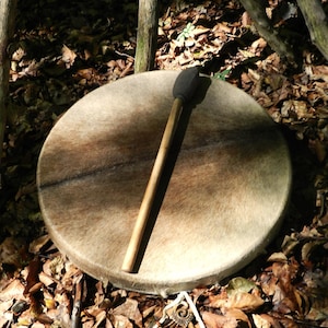 Pode incluir: Um tambor de moldura redondo com uma pele animal texturizada castanho-clara e uma baqueta de madeira pousada em cima. O tambor apresenta uma moldura circular de madeira e um pequeno pendente decorativo de metal preso à borda com um cordão branco.